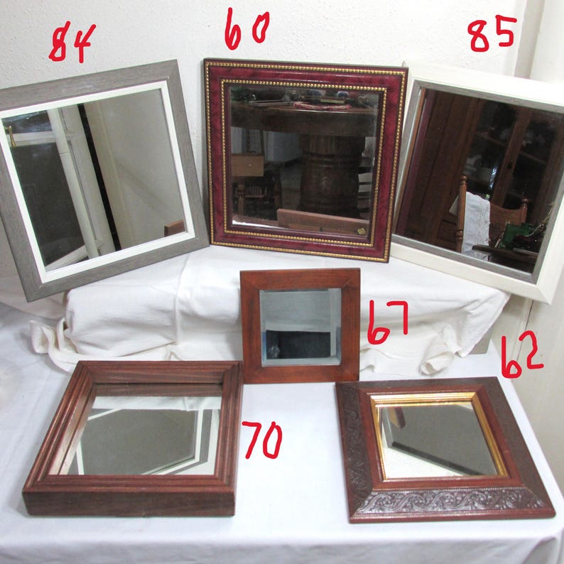 May include: A selection of square mirrors with diverse frame designs. The frames feature a grey wood effect, red and gold detailing, a white frame, dark wood, and a decorative brown frame with gold trim. The mirrors are arranged on a white surface.