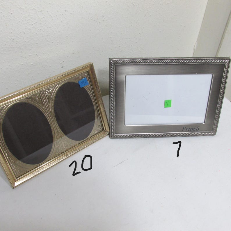 May include: Two picture frames. One is gold-toned with two oval openings. The other is silver-toned with a rectangular opening and the word "Friends" at the bottom. Both frames are empty.