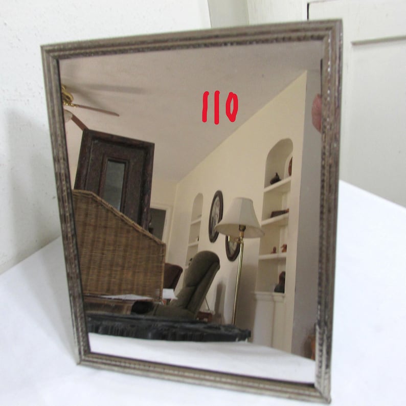 May include: A rectangular mirror with a silver-coloured frame. The mirror reflects a room with a wicker chair, a lamp, and a ceiling fan. The number "110" is written in red on the mirror's surface.