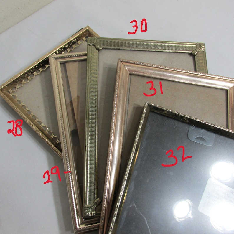 May include: A collection of gold-toned picture frames in various styles. The frames have decorative edges and are empty, ready for photos. The sizes vary, with some having ornate detailing and others a more simple design.