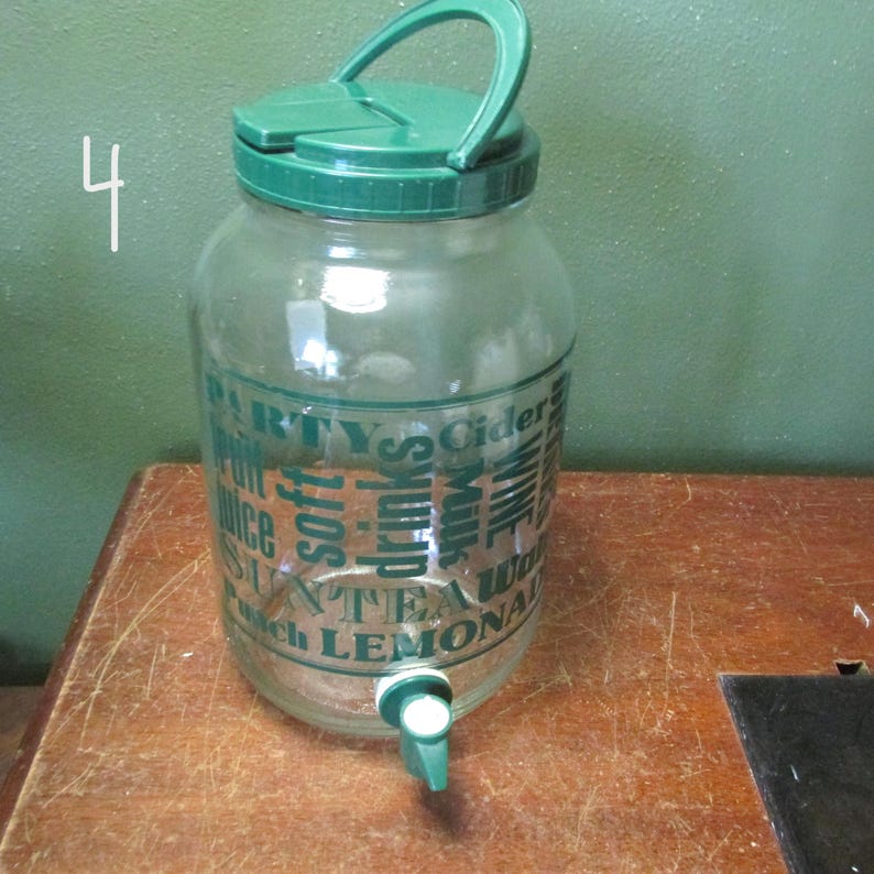 May include: A clear glass beverage dispenser with a green plastic lid featuring a handle and a green spigot at the base. The glass body is printed with green text including the words PARTY, CIDER, JUICE, SOFT DRINKS, MILK, WINE, SUN TEA, PUNCH, and LEMONADE.