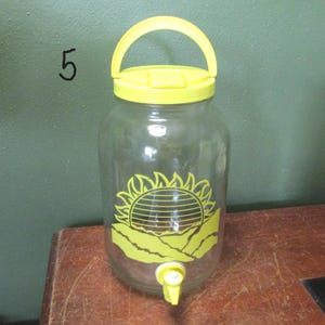 May include: A clear glass beverage dispenser featuring a bright yellow plastic lid with an integrated handle and a matching yellow spigot at the base. The front of the glass jar displays a yellow graphic of a sun rising over mountain peaks.