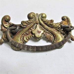 May include: Ornate antique drawer pull with a gold finish and a dark brown handle. The decorative hardware features a detailed, scrolling design with a central floral element and ring attachments.