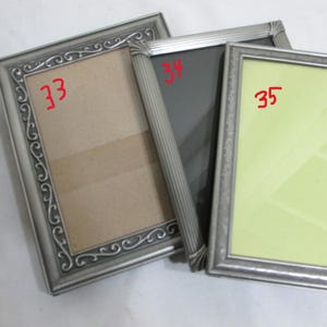 May include: Three silver-colored picture frames with ornate detailing. The frames have different colored backgrounds: beige, black, and light green. The frames are arranged at slightly different angles, showcasing their design.