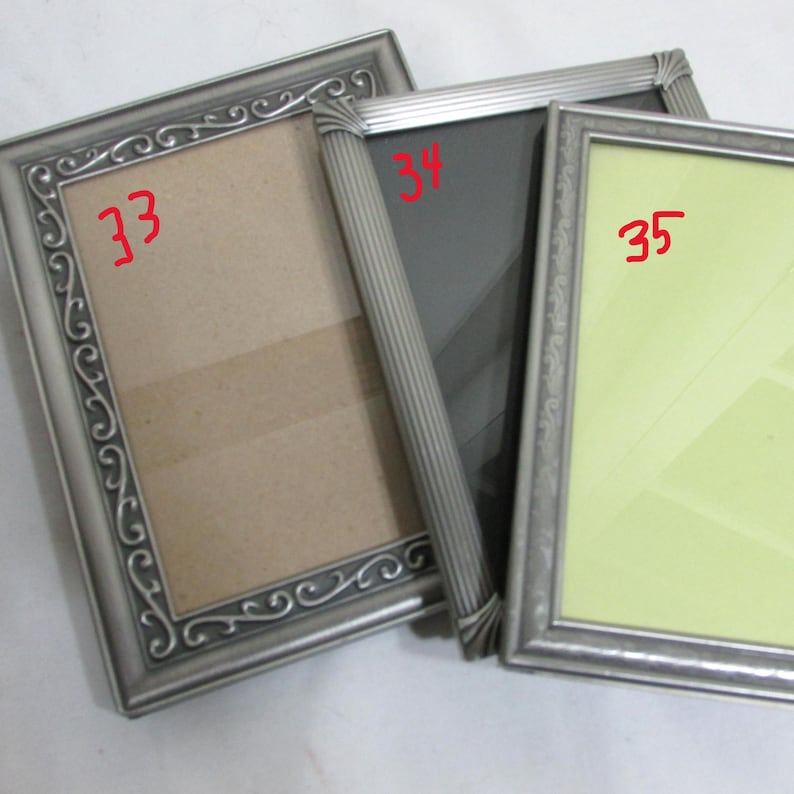 May include: Three silver-colored picture frames with ornate detailing. The frames have different colored backgrounds: beige, black, and light green. The frames are arranged at slightly different angles, showcasing their design.