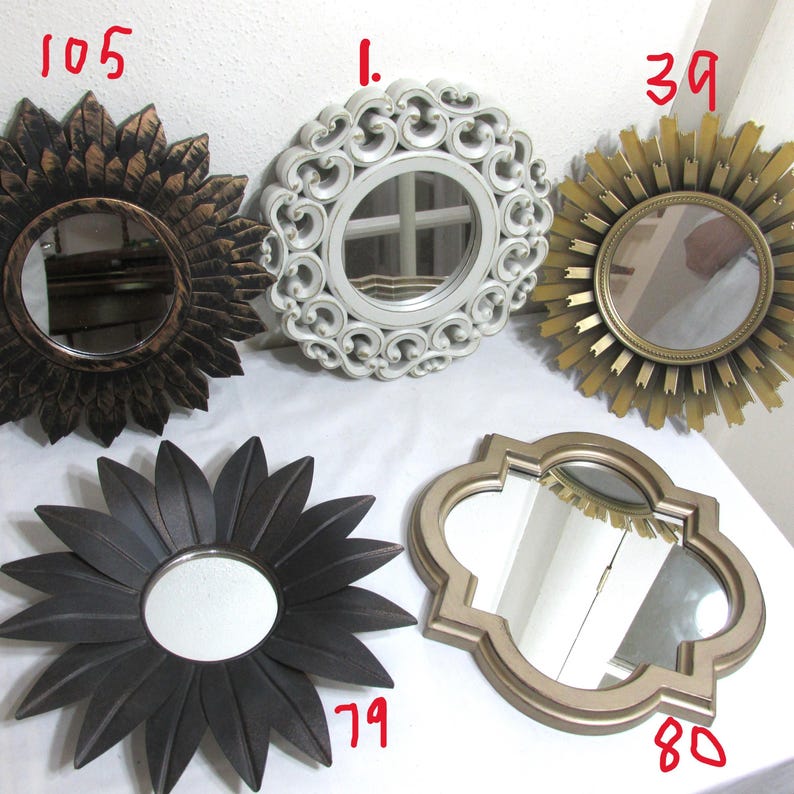 May include: Five decorative mirrors in various shapes and styles. Includes a sunflower-shaped mirror, a white ornate mirror, a sunburst mirror, a flower-shaped mirror, and a gold quatrefoil mirror. Each mirror has a reflective surface.