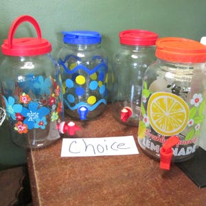 May include: Four clear glass beverage dispensers with spigots and lids. One has a red lid and blue floral print, one has a blue lid and yellow polka dots, one has a red lid, and one has an orange lid with a lemon graphic and the text Freshly Squeezed Lemonade.