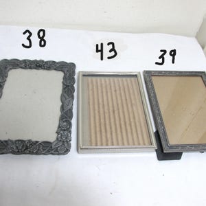 May include: Three vintage picture frames. The frames are in various styles, including a dark gray frame with floral detailing, a silver frame with a beaded edge, and a dark gray frame with a decorative border. Each frame is empty.
