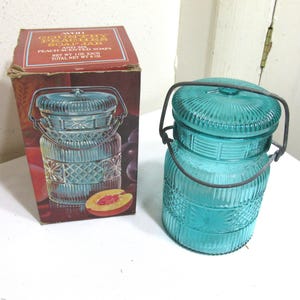 May include: A vintage, turquoise-colored glass jar with a metal handle and lid, next to a matching box. The box features an illustration of the jar and text that reads "Avon Country Peach Soap Jar". The jar has a decorative, textured design.
