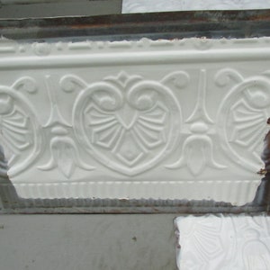 May include: White decorative tin ceiling tile with an embossed floral and geometric pattern. The design features heart-shaped elements, stylized leaves, and symmetrical details. The tile has a textured surface and a slightly distressed appearance.