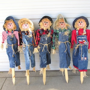 May include: Five scarecrows with straw hair and smiling faces. They wear denim overalls, plaid or striped shirts, and hats. The scarecrows are attached to wooden stakes, perfect for outdoor fall decor. One has a red shirt with "FIRE DEPT" on the overalls.