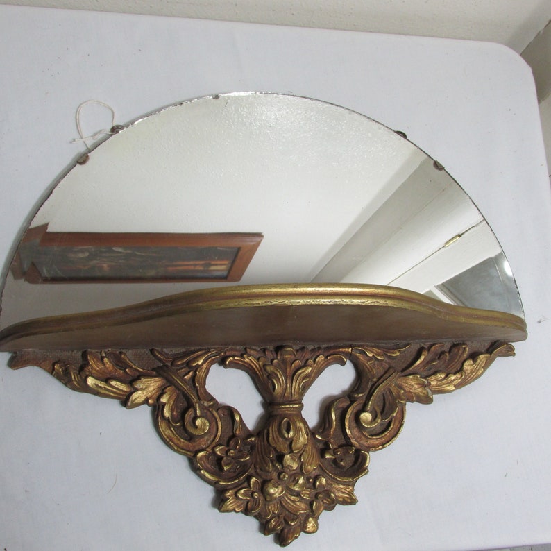 May include: A gold-painted wooden shelf with a half-round mirror and ornate scrollwork. The shelf is designed to be mounted on a wall.