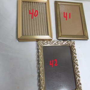 May include: Three vintage picture frames with gold-colored metal frames. One frame has a corrugated cardboard backing, another has a tan backing, and the third has a dark backing. Each frame has a red number on it.