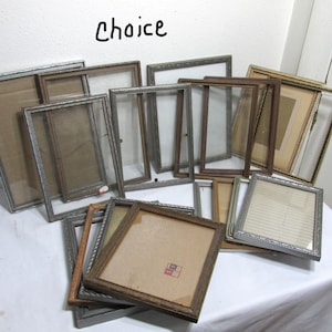 May include: A collection of vintage picture frames in various sizes and styles. The frames are made of wood and metal with silver and gold accents. Some frames have glass panes, while others have cardboard backing. The frames are arranged in a pile on a white surface.