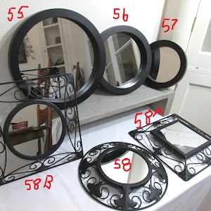 May include: A selection of black-framed mirrors in different shapes and sizes. Includes round, square, and decorative designs. The mirrors are displayed on a white surface and against a white wall.