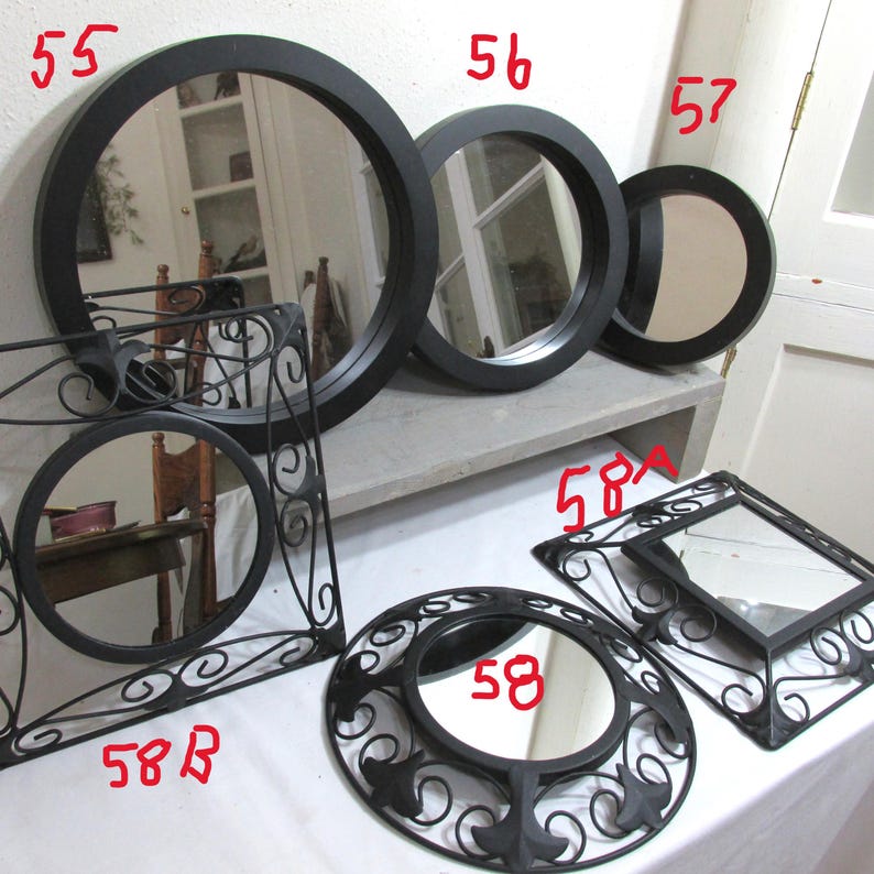 May include: A selection of black-framed mirrors in different shapes and sizes. Includes round, square, and decorative designs. The mirrors are displayed on a white surface and against a white wall.