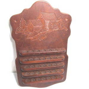 May include: A wall-mounted wooden display rack with a medium brown finish, featuring a carved relief of a water mill and cabin at the top. The unit includes five horizontal shelves, each with a series of small circular indentations for storage.