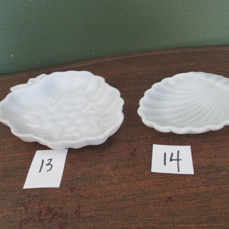 May include: Two white, decorative dishes. One dish is shaped like a bunch of grapes, and the other is shaped like a seashell. Each dish has a number on a small white tag: 13 and 14.