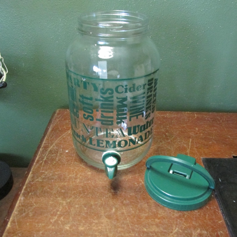 May include: A clear glass beverage dispenser with a green plastic spigot and a matching green lid. The exterior features dark green text listing various drink types including Cider, Wine, Water, Lemonade, Tea, Milk, and soft drinks in a collage.