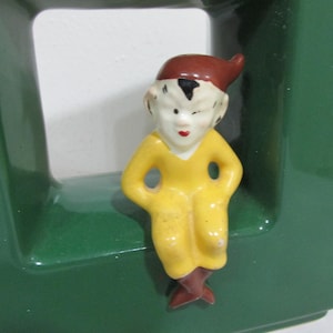 May include: A small, vintage ceramic figurine of a seated elf. The elf wears a yellow outfit, brown shoes, and a brown pointed hat. The figurine has a white face with red lips and black hair. The elf is positioned on a green surface.