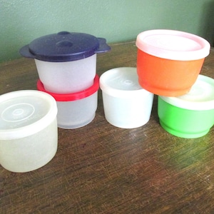 May include: Five plastic containers with lids. The containers are in various colors, including white, orange, green, and clear. The lids are white, pink, and blue.