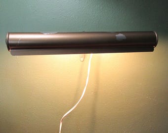 May include: A bronze-colored picture light fixture with a rectangular shape. The light is mounted on a wall and has a visible power cord. The light is turned on, illuminating the wall below.
