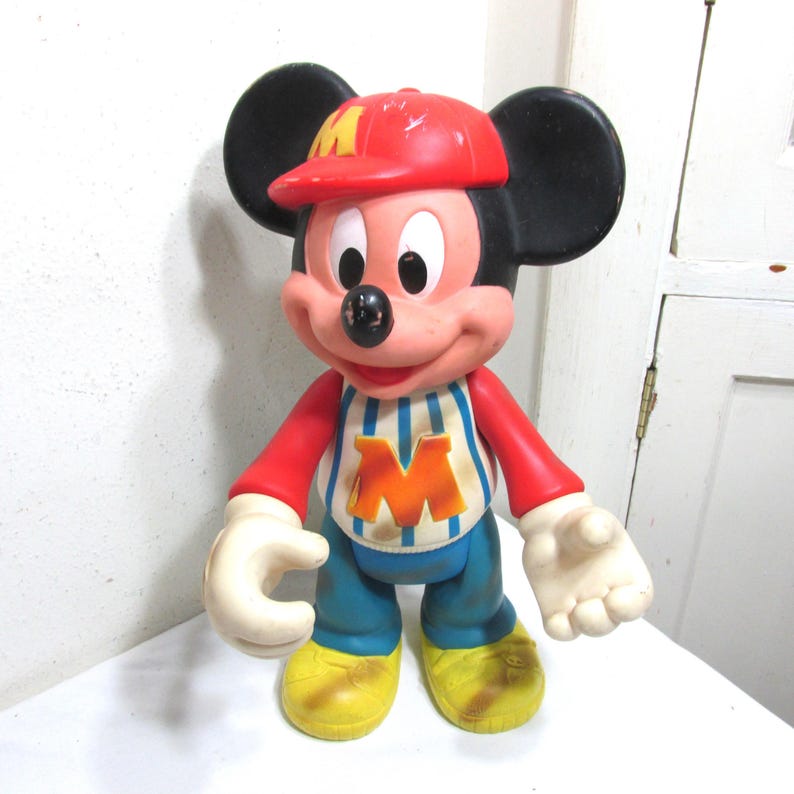 K&ouml;nnte beinhalten: Eine Vintage-Mickey-Mouse-Figur. Die Figur tr&auml;gt eine rote Kappe, ein rotes Langarmshirt, eine blaue Hose und gelbe Schuhe. Das Hemd hat ein blau-wei&szlig; gestreiftes Muster mit einem orangefarbenen "M" auf der Vorderseite. Die Figur hat schwarze Ohren und wei&szlig;e Handschuhe.