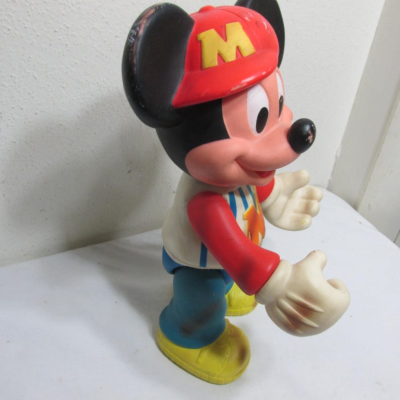 K&ouml;nnte beinhalten: Eine Vintage-Mickey-Mouse-Figur. Die Figur tr&auml;gt eine rote Baseballkappe mit einem gelben "M", ein wei&szlig;es Hemd mit roten &Auml;rmeln, eine blaue Hose und gelbe Schuhe. Die Figur hat wei&szlig;e Handschuhe und einen l&auml;chelnden Gesichtsausdruck.