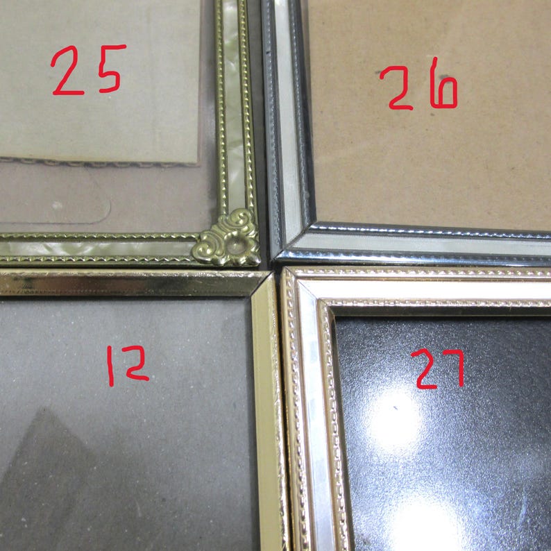 May include: A collection of four picture frames with different styles and colors. The frames are numbered 12, 25, 26, and 27 in red. The frames have gold, silver, and pearl-like finishes. The frames are empty, ready to display photos or artwork.