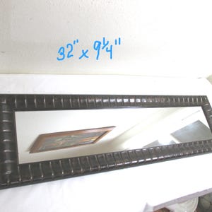 May include: Rectangular mirror with a dark brown, textured frame. The mirror measures 32 inches by 9.75 inches. The frame has a repeating pattern of vertical lines. The mirror reflects a painting and a white wall.