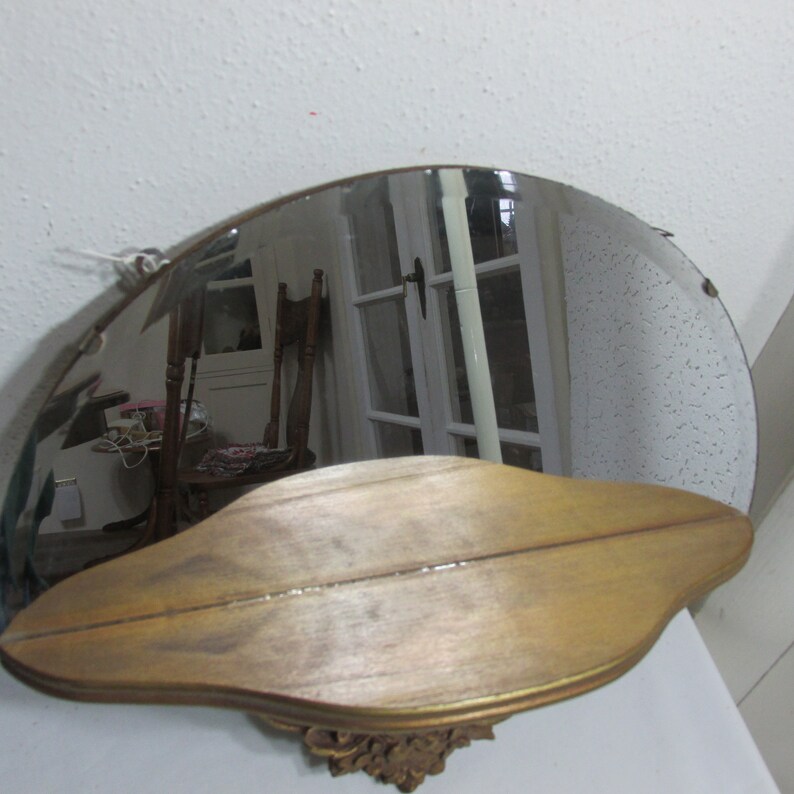 May include: A vintage, gold-toned, half-round wall mirror with a shelf underneath. The mirror has a beveled edge and the shelf is curved to match the shape of the mirror.
