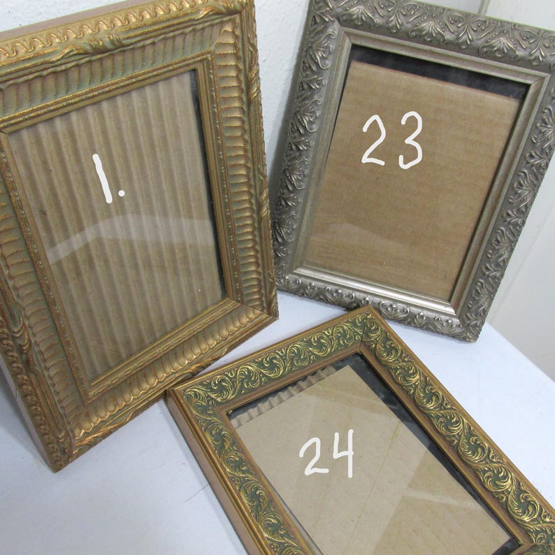 May include: Three ornate picture frames in gold and silver tones. One frame has the number "1" written on the glass, another has "23", and the third has "24". The frames have decorative edges and are empty.