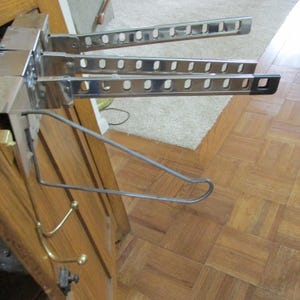 May include: A wall-mounted metal garment rack featuring three horizontal chrome-plated arms with evenly spaced rectangular slots for hangers. Below the arms, a single wire hanger hook and a brass-toned double hook are attached to the base.
