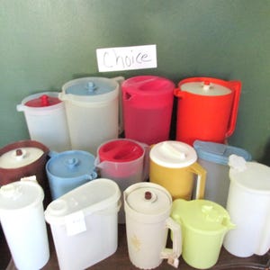 May include: A collection of 12 vintage plastic pitchers in various colors, including red, blue, yellow, white, and brown. The pitchers are arranged in a pyramid shape on a wooden surface. The word "Choice" is written on a white piece of paper in the background.