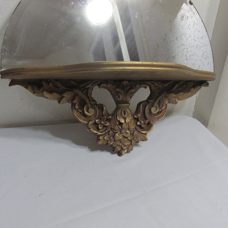 May include: A gold-painted, ornate, half-round wall mirror with a shelf. The mirror has a scalloped edge and the shelf is supported by decorative, scroll-like brackets.