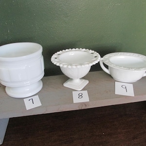 May include: Three white decorative ceramic pieces. The pieces include a small pedestal bowl with a scalloped rim, a small handled bowl, and a cylindrical container. Each piece is numbered with a small white tag.