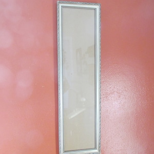 May include: A long, rectangular picture frame with a silver-colored, ornate border. The frame is empty, with a clear glass pane. The frame is displayed against a solid, reddish-orange wall.