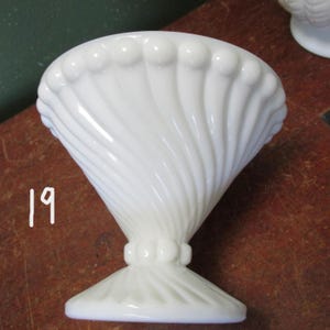 May include: A white, vintage-style pedestal bowl with a flared, ribbed design. The bowl has a wide, scalloped rim and a decorative base. The number "19" is written in white on the side of the bowl. The bowl is likely made of glass or ceramic.