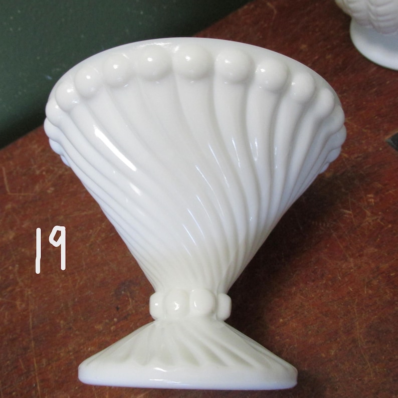May include: A white, vintage-style pedestal bowl with a flared, ribbed design. The bowl has a wide, scalloped rim and a decorative base. The number "19" is written in white on the side of the bowl. The bowl is likely made of glass or ceramic.