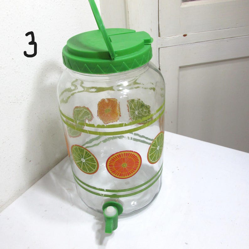 May include: A clear glass beverage dispenser featuring a green plastic lid with a handle and a green spigot at the base. The exterior of the glass is decorated with a repeating pattern of orange and lime slices, accented by horizontal green lines.