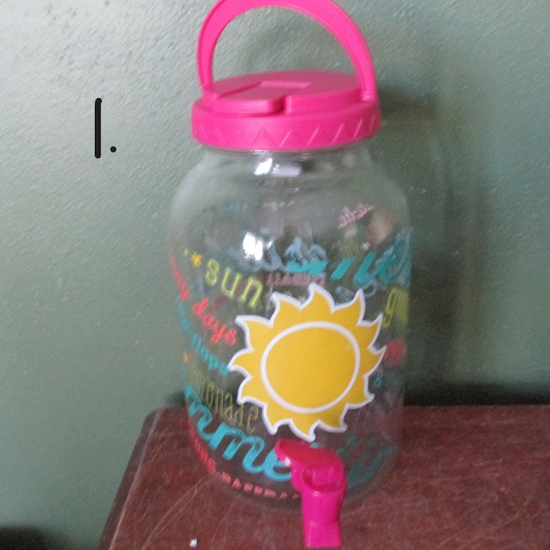 May include: A clear glass beverage dispenser with a bright pink plastic lid featuring a handle and a matching pink spigot at the base. The exterior is decorated with a large yellow sun graphic and various words including sun, days, and lemonade.