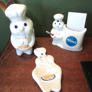 May include: Three white ceramic Pillsbury Doughboy kitchen items including a standing utensil holder, a flour sack shaped note holder with a blue Pillsbury logo, and a flat spoon rest, all featuring the iconic character with blue eyes and a chef hat.