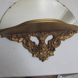 May include: A gold-painted wooden shelf with ornate scrollwork and a half-circle mirror. The shelf is designed to be mounted on a wall.