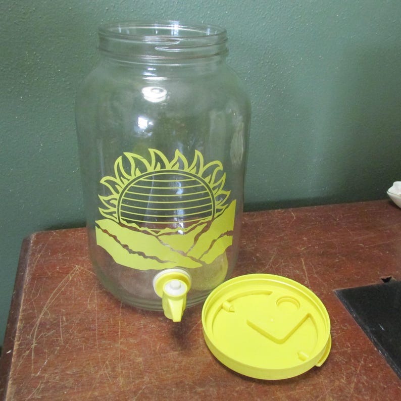 May include: A clear glass beverage dispenser featuring a yellow graphic of a sun rising over mountains on the front. The container includes a yellow plastic spigot at the base and a matching yellow circular lid placed beside the glass vessel.