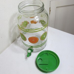 May include: A clear glass beverage dispenser featuring a printed pattern of green lime and orange citrus slices, accented by horizontal green stripes. The dispenser includes a green plastic spigot at the base and a matching green plastic lid.