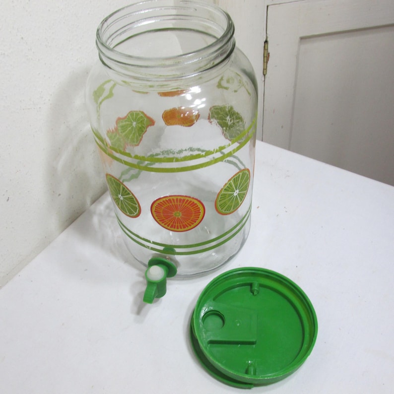 May include: A clear glass beverage dispenser featuring a printed pattern of green lime and orange citrus slices, accented by horizontal green stripes. The dispenser includes a green plastic spigot at the base and a matching green plastic lid.