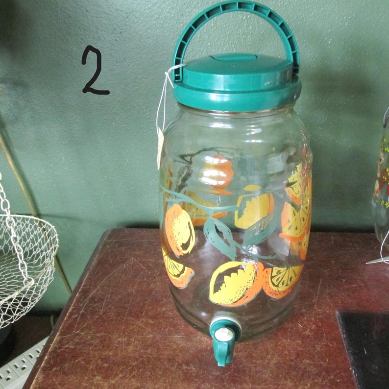 May include: A clear glass beverage dispenser featuring a green plastic lid with a handle and a green plastic spigot at the base. The glass body is decorated with a painted pattern of orange and yellow citrus fruit slices and green leaves.