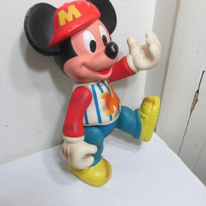 K&ouml;nnte beinhalten: Vintage Mickey Mouse Figur in Gehposition. Die Figur tr&auml;gt eine rote Kappe mit einem gelben "M", ein gestreiftes Hemd, blaue Hosen und gelbe Schuhe. Die Figur hat schwarze Ohren, ein rosa Gesicht und wei&szlig;e Handschuhe.