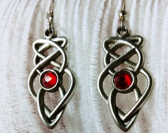 May include: A pair of silver-toned earrings with a Celtic knot design. Each earring features a red, oval-shaped gemstone at the center. The earrings hang from a simple hook and are displayed against a white, feathery background.