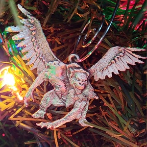 May include: A silver-colored Christmas ornament depicting a gargoyle with wings. The ornament is hanging from a Christmas tree with colorful tinsel.